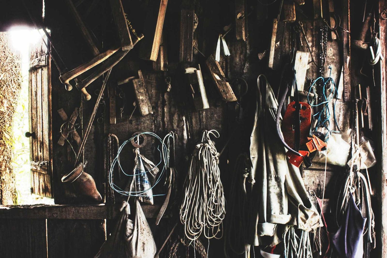 Weathered workshop wall covered in hanging hand tools, coiled rope and worn work clothes
