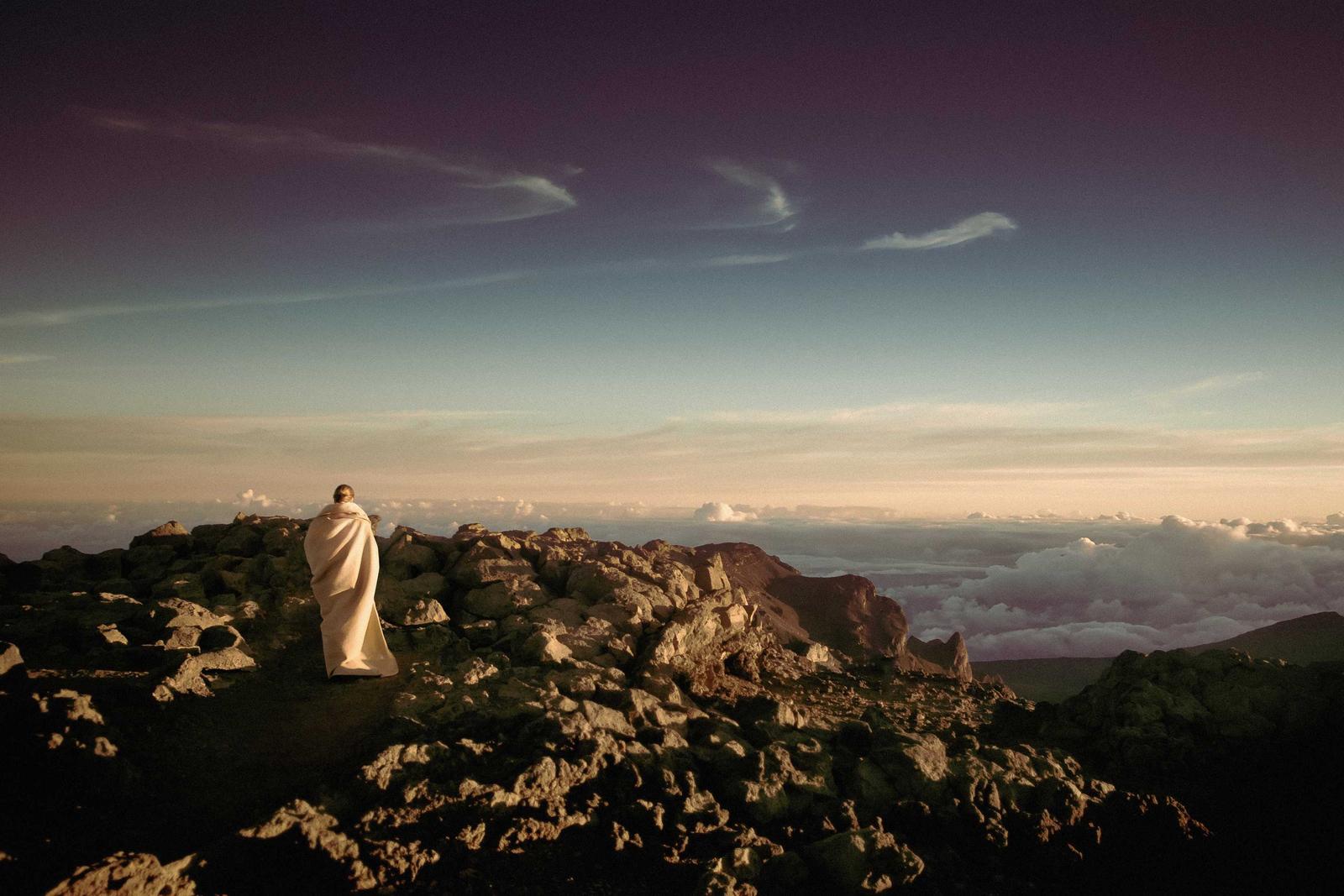 Figure wrapped in flowing white cloth standing on dark volcanic rock above a sea of clouds at dusk