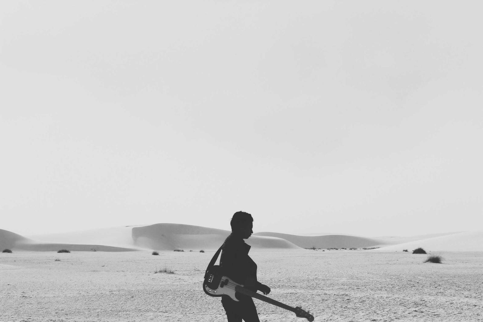 Silhouette of a person walking through pale dunes carrying a bass guitar, black and white